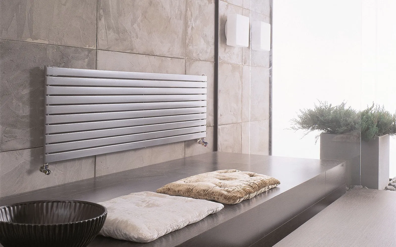 Ein flacher, silberner Heizkörper ist an einer strukturierten, grauen Steinwand montiert. 