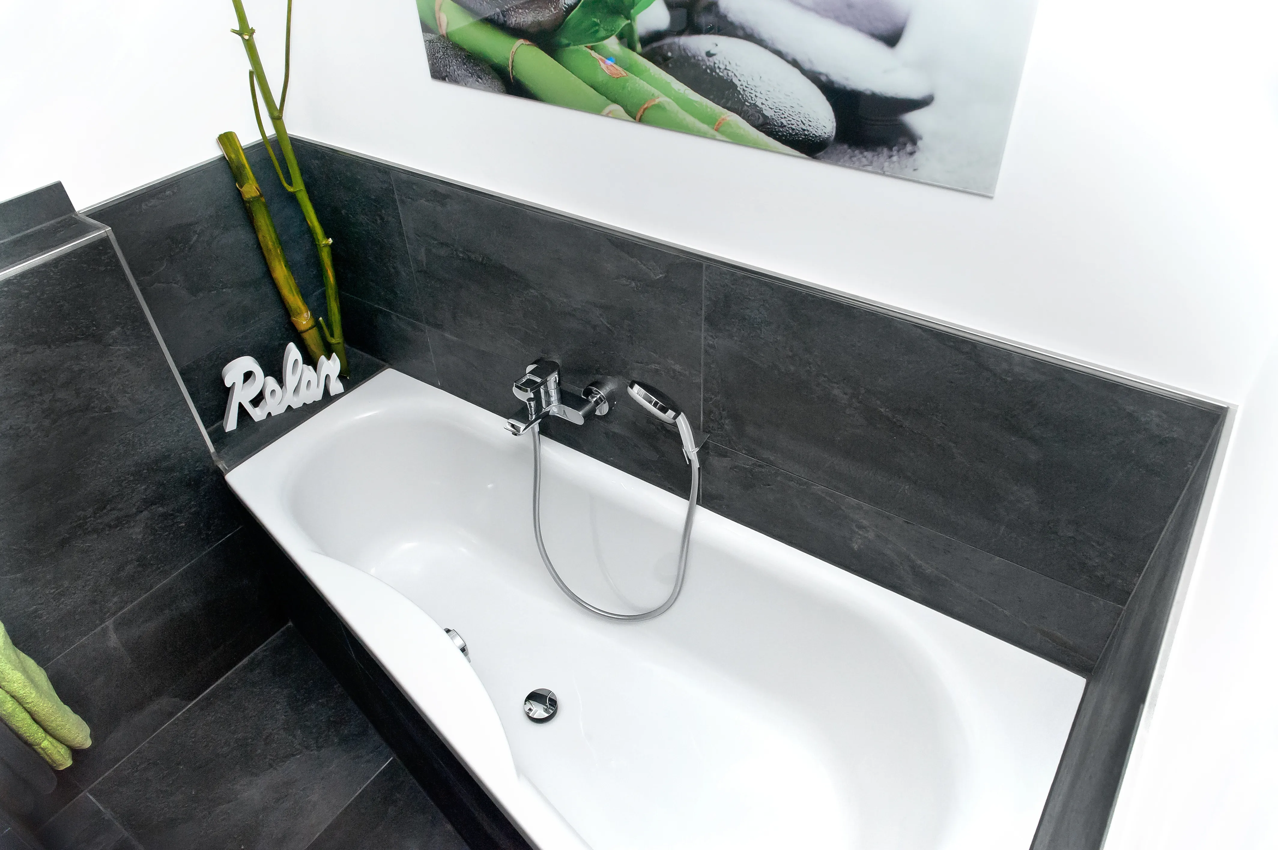 Asymetrische Badewanne mit grauen Fliesen und Schriftzug "Relax" als Deko. 