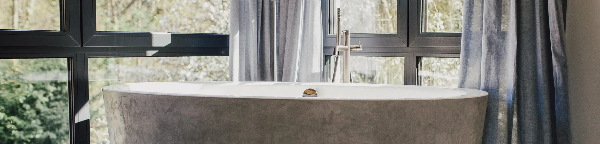 Minimalistische Badewanne mit edler Edelstahlarmatur vor einer raumhohen Fensterfront mit halbtransparenten Vorhängen.