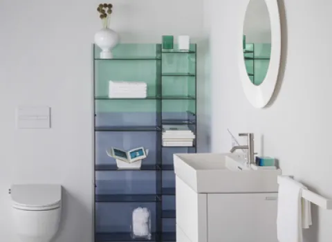 Ein helles Badezimmer mit einer wandhängenden Toilette und einem weißen Waschtisch. Ein rundes, weiß gerahmtes Spiegel hängt über dem Waschbecken, daneben ein Handtuchhalter.