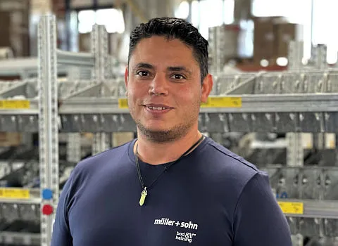 Ein junger Anlagenmechaniker mit T-Shirt von müller + sohn steht in einer Lagerhalle.