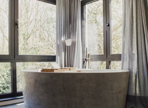 Eine freistehende Badewanne mit mattem Stein-Finish steht vor großen Panoramafenstern, durch die ein Blick ins grüne Waldstück fällt. 