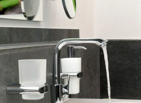 Waschtisch mit Armatur von Keuco vor grauer Fliesenwand und Wandspiegel mit Kosmetikspiegel. 