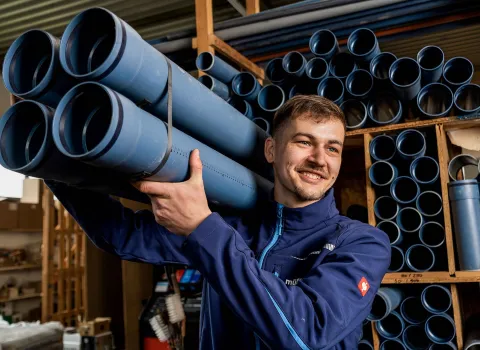Ein junger Anlagenmechaniker SHK trägt mehrere blaue Kunststoffrohre auf der Schulter und lächelt dabei.