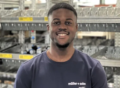 Lagerarbeiter der Firma müller+sohn steht vor einem Regal und lächelt in die Kamera.